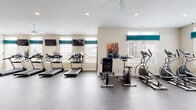 Located at 4900 Gender Rd in Columbus, Ohio, this single-floor fitness center is a well-equipped community amenity designed to support a comprehensive workout routine. The space is bright and open, with neutral beige walls accented by teal window valances, recessed lighting, and ceiling fans that contribute to a clean, comfortable environment.

The cardio zone is a standout feature, housing multiple treadmills with individual digital displays, an elliptical trainer, and a stationary rowing machine — all positioned along windowed walls to allow natural light and outdoor views during workouts. Wall-mounted flat-screen TVs are positioned throughout this area for added convenience.

A dedicated strength training section features a full rack of dumbbells in varying weights, an adjustable weight bench, medicine balls, and a multi-station cable machine, providing versatility for users of all fitness levels. Full-length mirrors line the walls, enhancing the sense of space and allowing users to monitor their form.

The durable rubber flooring throughout provides excellent traction and cushioning, while a separate free-weight alcove with large interior windows overlooks an adjacent lounge or clubhouse area, offering an integrated community feel. The layout is thoughtfully organized, providing ample open floor space between equipment zones for safe and comfortable movement.