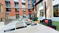 This single-floor space offers a compelling blend of outdoor amenity and interior utility. The standout feature is a well-appointed outdoor courtyard, enclosed by a multi-story urban building facade of warm brick and glass. The courtyard is designed for relaxed gathering, furnished with contemporary wicker and upholstered lounge chairs accented with colorful throw pillows. A central raised planter with mature greenery anchors the space, while a freestanding fireplace adds a focal point and warmth. Light-toned deck and paver surfaces extend the seating area, creating a clean, modern aesthetic. Additional bench seating with red cushions lines one side, offering versatile arrangements for socializing.

The interior includes a bathroom finished in a straightforward, functional style. Clean white walls and neutral-toned countertops provide a simple backdrop, while a large wall mirror above the sink enhances the sense of openness. Chrome hardware and a square-profile towel bar add modest modern touches. The bathroom is equipped with a standard vanity, toilet, and a shower/tub combination featuring a light blue shower curtain with a subtle geometric pattern. Adequate overhead ventilation and lighting complete the practical layout. Together, the outdoor and indoor elements create a space suited for both community gathering and everyday convenience.