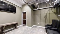 Located at 390 Capitol Ave, this single-floor space offers a thoughtfully designed interior that combines industrial aesthetics with modern finishes. The space features sage-green walls that create a calm, professional atmosphere, complemented by light-toned, patterned carpet tiles laid in a geometric arrangement that adds visual interest underfoot. Exposed ductwork and conduit runs along the ceiling, lending an industrial character to the space while maintaining a clean, polished look. The ceiling also incorporates recessed and track lighting, ensuring the area is well-lit throughout. A dark wood-framed glass door serves as a functional and attractive entry point, adding warmth against the neutral color palette. The space includes built-in wall-mounted display screens, making it well-suited for presentations or media use. Simple, rustic furnishings — including a wooden console table — contribute to a balanced blend of raw and refined design. White baseboards provide a crisp contrast to the muted wall tones, tying the overall aesthetic together. The open layout and adaptable features make this space suitable for a variety of commercial or professional uses.