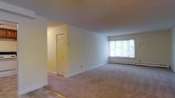 This single-floor space presents a clean, open layout centered around a spacious living area finished with wall-to-wall neutral carpeting that extends throughout. The room is bathed in natural light streaming through a large multi-pane window along the far wall, offering views of lush greenery outside. Smooth, white walls and white trim create a bright, airy atmosphere, while the generous proportions of the main living area allow for flexible furniture arrangements. A baseboard heating unit runs along the exterior wall beneath the window, providing an efficient heating solution. A recessed entryway with a classic six-panel white door is visible off the main living space, suggesting a closet or secondary room that adds functional storage or additional utility to the layout. The transition area near the entry features a hard flooring threshold, contrasting with the soft carpeting of the main living zone. Overall, the space maintains a straightforward, well-lit, and practical character with neutral finishes that provide a versatile foundation for personalization.