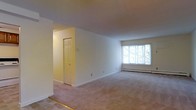 This single-floor space presents a clean, open layout centered around a spacious living area finished with wall-to-wall neutral carpeting that extends throughout. The room is bathed in natural light streaming through a large multi-pane window along the far wall, offering views of lush greenery outside. Smooth, white walls and white trim create a bright, airy atmosphere, while the generous proportions of the main living area allow for flexible furniture arrangements. A baseboard heating unit runs along the exterior wall beneath the window, providing an efficient heating solution. A recessed entryway with a classic six-panel white door is visible off the main living space, suggesting a closet or secondary room that adds functional storage or additional utility to the layout. The transition area near the entry features a hard flooring threshold, contrasting with the soft carpeting of the main living zone. Overall, the space maintains a straightforward, well-lit, and practical character with neutral finishes that provide a versatile foundation for personalization.