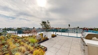 Located in the heart of Venice, Los Angeles, this rooftop terrace at 1503 E Venice Blvd offers an expansive outdoor amenity space designed for both relaxation and social gatherings. The terrace is finished with large-format porcelain tiles that extend across the open deck, creating a clean, modern aesthetic throughout.

Lush, drought-tolerant landscaping is thoughtfully integrated into raised garden beds and white cubic planters, featuring ornamental grasses, native shrubs, and young trees that add a natural, organic contrast to the urban setting. Accent lighting installed within the garden beds allows the greenery to be enjoyed well into the evening.

Multiple seating areas are distributed across the terrace, including bar-height dining tables with woven chairs and loungers shaded by bold black-and-white striped market umbrellas. A stainless steel BBQ grill station provides a dedicated outdoor cooking area, making the space ideal for entertaining.

The terrace is enclosed by a distinctive bright blue metal railing system that adds a pop of color against the white structural elements and open sky. Panoramic views of the surrounding Venice and Los Angeles neighborhoods — including palm trees and low-rise architecture — are visible from all sides of the deck, offering a quintessential Southern California backdrop to this well-appointed rooftop retreat.