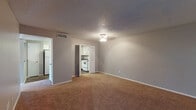 This single-story residence offers a straightforward, functional layout with neutral gray walls and textured ceilings throughout. The living area is carpeted in warm brown tones and receives natural light through a large window flanking the front entry door. A ceiling fan provides overhead lighting and ventilation. The space opens toward a kitchen and additional rooms, creating an accessible flow through the unit.

The kitchen is well-equipped with white shaker-style cabinetry extending to the upper walls, providing ample storage. Granite-style countertops run along the perimeter, complemented by a double stainless steel sink, a white electric range, and a dishwasher. Dark hardwood-look flooring anchors the kitchen area.

The bedroom features plush carpeting, a ceiling fan with light fixture, a standard closet, and a window that allows natural light and outdoor views. Direct access to a bathroom is conveniently located within the bedroom.

Two bathrooms are present, each featuring white vanity cabinetry with granite-style countertops, large mirrors, and dark wood-look flooring. One bathroom includes a tall linen storage cabinet and grab bar, adding practical utility to the space.