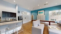 Located at 1767 Hermitage Blvd in Tallahassee, Florida, this single-floor community amenity space offers a thoughtfully designed environment for gathering, relaxing, and handling everyday conveniences. The interior is defined by soaring vaulted ceilings, warm hardwood-style flooring, and a bright, modern color palette featuring soft blues, crisp white trim, and a bold teal accent wall that anchors the space with a pop of color.

The lounge area is generously furnished with plush grey swivel chairs, a sofa, and an upholstered ottoman centered around a coffee table. A wall-mounted flat-screen TV and a sleek electric fireplace add to the comfort of this gathering zone. Expansive multi-pane windows flood the space with natural light and frame views of the lush, landscaped outdoor pool area.

A kitchenette features granite countertops, dark espresso cabinetry, a sink, microwave, coffee maker, and a spacious bar-height counter with barstool seating — ideal for casual use. Package management lockers and additional storage units are conveniently integrated into the space. A shuffleboard table adds a recreational element, rounding out the amenities. Recessed lighting throughout ensures a clean, polished aesthetic. Glass partition doors connect adjacent areas, enhancing the open, airy feel of the overall layout.