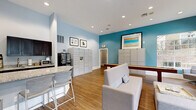 Located at 1767 Hermitage Blvd in Tallahassee, Florida, this single-floor community amenity space offers a thoughtfully designed environment for gathering, relaxing, and handling everyday conveniences. The interior is defined by soaring vaulted ceilings, warm hardwood-style flooring, and a bright, modern color palette featuring soft blues, crisp white trim, and a bold teal accent wall that anchors the space with a pop of color.

The lounge area is generously furnished with plush grey swivel chairs, a sofa, and an upholstered ottoman centered around a coffee table. A wall-mounted flat-screen TV and a sleek electric fireplace add to the comfort of this gathering zone. Expansive multi-pane windows flood the space with natural light and frame views of the lush, landscaped outdoor pool area.

A kitchenette features granite countertops, dark espresso cabinetry, a sink, microwave, coffee maker, and a spacious bar-height counter with barstool seating — ideal for casual use. Package management lockers and additional storage units are conveniently integrated into the space. A shuffleboard table adds a recreational element, rounding out the amenities. Recessed lighting throughout ensures a clean, polished aesthetic. Glass partition doors connect adjacent areas, enhancing the open, airy feel of the overall layout.