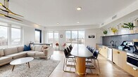 Located at 2703 Girard Ave S in Minneapolis, MN, this single-floor space seamlessly combines a kitchen and living area into one well-designed, open-concept layout. Large floor-to-ceiling windows wrap around the space, flooding the interior with abundant natural light and offering sweeping views of the surrounding Minneapolis skyline and neighborhood rooftops.

The living area is thoughtfully appointed with warm hardwood flooring, a starburst pendant light fixture, a wall-mounted flat-screen display, and a comfortable seating arrangement centered around a marble-topped coffee table. The space accommodates multiple seating zones, making it ideal for both relaxed gatherings and more structured meetings.

The kitchen features sleek, light wood cabinetry with gold hardware accents, a bold grey stone countertop that extends into a generous curved island, a textured mosaic tile backsplash, and stainless steel appliances including a full-size refrigerator. Wall-mounted sconces with potted greenery add a fresh, welcoming touch above the counters.

An oval dining table with seating for several occupants bridges the kitchen and living zones. Direct access to a furnished outdoor terrace extends the usable space further, offering open-air seating with panoramic views of the Minneapolis cityscape — perfect for outdoor gatherings or a breath of fresh air.