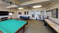 Located at 3304 E 44th Ave in Spokane, Washington, this single-floor community game room is designed for recreation and social interaction. The space is generously proportioned, with a layout that comfortably accommodates multiple gaming stations simultaneously. At its center sits a full-size pool table with a vibrant green felt surface, complemented by a long shuffleboard table along one wall and an air hockey table, providing a well-rounded selection of classic games. Overhead, flush-mounted fluorescent lighting illuminates the carpeted floor evenly throughout the room. A dedicated lounge nook features a high-top bistro table with bar-height chairs, positioned beside large vertical-blind windows that invite ample natural light and offer views of the surrounding exterior. On the opposite end of the room, a wall-mounted flat-screen television is flanked by framed artwork with an automotive theme, and a guitar on a stand adds a creative touch to the atmosphere. Additional décor, including plaques and small framed pieces, contributes to the room's personalized character. The space benefits from direct exterior access through a glass-panel door, further enhancing connectivity to the outdoors. Overall, the game room presents a welcoming, activity-rich environment suited for community gatherings and leisure.