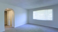 This single-story space offers a straightforward and functional layout characterized by clean, neutral design elements throughout. The main living area is finished with wall-to-wall carpet in a neutral tone, providing a soft underfoot feel that complements the light-colored walls. A textured popcorn ceiling adds dimension overhead, while white baseboards frame the perimeter of the room cleanly.

A notable architectural detail is the arched doorway along one wall, lending a subtle decorative touch that breaks the uniformity of the otherwise simple layout. This curved archway transitions into an adjacent area, hinting at a connected yet distinct space within the floor plan.

Natural light enters through a large, multi-pane window fitted with horizontal blinds, offering both brightness and privacy control. The window provides a view of exterior greenery, adding a sense of connection to the outdoors. The overall ambiance of the space is bright, open, and neutral, making it a versatile canvas well-suited for a variety of furnishing styles and personal preferences. The single-floor configuration ensures ease of movement throughout the space.