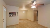 This single-floor space offers a straightforward, functional layout centered around an open living and dining area. The main living space is finished with wall-to-wall carpeting and neutral-toned walls that create a warm, understated atmosphere. A ceiling fan with an integrated light fixture is centrally positioned overhead, providing both airflow and ambient lighting. A pendant light fixture above the dining area adds a focused point of illumination, helping to define the space.

A pass-through window connects the kitchen to the living and dining area, offering a sense of openness while maintaining a visual separation between the two spaces. The kitchen, visible through the pass-through, features dark wood cabinetry and includes appliances such as a microwave. The transition from the tiled kitchen flooring to the carpeted living area is clearly delineated, emphasizing the distinct functional zones.

Toward the rear of the space, a hallway leads to additional rooms, with a bi-fold door visible, likely concealing a closet. The overall layout is practical and efficiently organized, making good use of the available floor space. The neutral color palette and consistent design elements throughout contribute to a cohesive and livable environment.