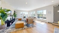 Located at 45 Gorge Rd E in Victoria, British Columbia, this single-floor space offers a thoughtfully designed open-plan layout that seamlessly integrates the kitchen and living areas into one cohesive, light-filled environment. Light oak hardwood flooring runs throughout, tying the space together and complementing the clean white walls and crisp recessed lighting overhead.

The living area is anchored by a wall of large picture windows and sliding glass doors that frame expansive outdoor views, flooding the interior with natural light. A warmly accented entryway features rich wood paneling, adding texture and contrast to the otherwise contemporary palette.

The kitchen is a standout highlight, appointed with sleek, flat-front cabinetry in a warm wood tone, stone countertops, a generous central island with bar seating for four, and stainless steel appliances. Statement pendant lights hang above the island, adding a modern, industrial-chic touch. A sliding glass door opens directly onto an outdoor terrace equipped for outdoor dining, making indoor-outdoor living effortless.

The space also accommodates a dining area with a solid wood table, offering a natural gathering point between the kitchen and living zones. Throughout, the design balances minimalist architecture with warm material choices, creating an inviting, functional, and bright environment ideal for both everyday living and entertaining.