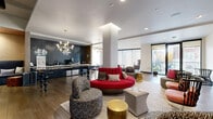 This single-floor communal lounge is thoughtfully designed to offer a dynamic, multi-functional gathering space with a contemporary aesthetic. The open layout is anchored by warm hardwood-style flooring and crisp white ceilings with recessed lighting, creating a bright and welcoming atmosphere throughout.

The main lounge area showcases an eclectic mix of seating, including a bold circular red sofa, patterned accent chairs, tufted ottomans, and gold cylindrical side tables, all arranged atop neutral area rugs. A large wall-mounted flat-screen television is positioned at one end, making the space equally suited for casual viewing or socializing.

Adjacent to the lounge is a well-appointed beverage bar featuring dark navy cabinetry with brass hardware, a glossy dark subway tile backsplash, and a striking marble-topped communal table with sculptural legs illuminated by modern gold chandeliers. A coffee station with a commercial-grade machine is neatly integrated into the counter.

A quieter alcove with floor-to-ceiling windows offers a sunlit retreat furnished with a curved sofa and tufted ottoman. Elsewhere, sliding glass doors open to an outdoor area, filling the space with natural light and providing a seamless indoor-outdoor connection. Structural columns punctuate the open floor plan while maintaining an airy, spacious feel.