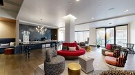 This single-floor communal lounge is thoughtfully designed to offer a dynamic, multi-functional gathering space with a contemporary aesthetic. The open layout is anchored by warm hardwood-style flooring and crisp white ceilings with recessed lighting, creating a bright and welcoming atmosphere throughout.

The main lounge area showcases an eclectic mix of seating, including a bold circular red sofa, patterned accent chairs, tufted ottomans, and gold cylindrical side tables, all arranged atop neutral area rugs. A large wall-mounted flat-screen television is positioned at one end, making the space equally suited for casual viewing or socializing.

Adjacent to the lounge is a well-appointed beverage bar featuring dark navy cabinetry with brass hardware, a glossy dark subway tile backsplash, and a striking marble-topped communal table with sculptural legs illuminated by modern gold chandeliers. A coffee station with a commercial-grade machine is neatly integrated into the counter.

A quieter alcove with floor-to-ceiling windows offers a sunlit retreat furnished with a curved sofa and tufted ottoman. Elsewhere, sliding glass doors open to an outdoor area, filling the space with natural light and providing a seamless indoor-outdoor connection. Structural columns punctuate the open floor plan while maintaining an airy, spacious feel.