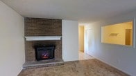 This single-story space offers a warm and functional layout with notable architectural character. The living area serves as the focal point of the home, anchored by a full-height brick accent wall housing an electric fireplace insert with a clean white mantel. The brick detailing adds a rustic, timeless quality to the space, creating a cozy atmosphere suitable for year-round enjoyment.

The flooring transitions from neutral-toned carpet in the living area to light tile in the adjacent hallway and connecting rooms, providing both comfort and practicality. The white walls throughout keep the interior feeling bright and open, complementing the natural texture of the brick wall.

A wide hallway extends from the living space, leading to additional rooms and suggesting a straightforward, accessible floor plan. A closet is visible along the hallway, offering convenient built-in storage. The overall color palette — warm beiges, whites, and brick tones — creates a cohesive and understated aesthetic throughout the single-level layout, making the space both livable and easy to personalize.