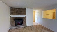 This single-story space offers a warm and functional layout with notable architectural character. The living area serves as the focal point of the home, anchored by a full-height brick accent wall housing an electric fireplace insert with a clean white mantel. The brick detailing adds a rustic, timeless quality to the space, creating a cozy atmosphere suitable for year-round enjoyment.

The flooring transitions from neutral-toned carpet in the living area to light tile in the adjacent hallway and connecting rooms, providing both comfort and practicality. The white walls throughout keep the interior feeling bright and open, complementing the natural texture of the brick wall.

A wide hallway extends from the living space, leading to additional rooms and suggesting a straightforward, accessible floor plan. A closet is visible along the hallway, offering convenient built-in storage. The overall color palette — warm beiges, whites, and brick tones — creates a cohesive and understated aesthetic throughout the single-level layout, making the space both livable and easy to personalize.