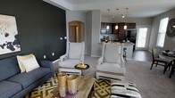 This single-floor residence offers a thoughtfully designed open-concept layout that seamlessly connects the living and kitchen areas, creating a spacious and inviting atmosphere. The living area is anchored by a bold dark accent wall, providing a striking contrast to the neutral gray tones found throughout the space. Plush carpeted flooring adds warmth underfoot, complemented by a patterned area rug in gold and black tones that adds visual interest. A pair of elegant high-back white accent chairs with nail-head trim detailing serve as a sophisticated focal point, paired with a rustic wood coffee table and coordinating decor pieces in gold tones. The kitchen is accessible via a stylish arched entryway and features dark espresso cabinetry, granite countertops, and a breakfast bar with barstool seating — ideal for casual dining. Pendant lighting in warm tones hangs above the kitchen island, adding an inviting glow. Stainless steel and black appliances complete the kitchen's modern aesthetic. Track lighting overhead provides ample illumination throughout the open space. The overall ambiance is contemporary and polished, blending bold design choices with comfortable, functional living spaces.