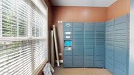 Located at 1660 Hampton Knoll Drive in Akron, Ohio, this dedicated package locker room serves as a secure and convenient parcel retrieval space. The entrance room is compact yet efficiently organized, finished with warm terracotta-toned walls and low-pile carpeted flooring throughout. The centerpiece of the space is a large Amazon Hub locker system, with banks of steel-blue lockers lining two walls from floor to ceiling, providing an extensive number of individual compartments for secure package storage. The locker system includes an interactive touchscreen terminal for easy package retrieval. Natural light filters into the room through large double-hung windows dressed with horizontal blinds, offering views of the surrounding greenery. A white paneled door with a glass insert provides access to an adjoining area, featuring a lever-style handle for ease of use. White baseboard trim provides a clean contrast against the warm wall color. The room is practical and purpose-built, designed to streamline package delivery and pickup for residents of the community.