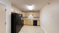 This single-floor space includes a well-equipped kitchen that serves as a central feature. The kitchen is outfitted with light wood cabinetry spanning both upper and lower sections, providing generous storage capacity. Black appliances, including a full-sized refrigerator, gas range with overhead microwave, and dishwasher, offer a practical contrast to the warm-toned cabinetry. The countertops run along an L-shaped layout, maximizing workspace efficiency. A double-basin sink is positioned beneath the upper cabinets along the back wall, adding convenience to the layout. The flooring features a neutral tile pattern that complements the overall palette of the space. A flush-mounted skylight fixture provides bright overhead lighting, keeping the kitchen well-lit. A baseboard heating unit runs along the base of the wall, ensuring warmth throughout the space. The overall aesthetic is clean and functional, with neutral tones creating a cohesive and understated ambiance suited for everyday living.
