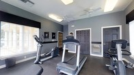 This fitness center offers a practical and well-maintained workout environment designed to meet everyday exercise needs. The space is finished with cool gray walls and dark flooring, creating a focused, energetic atmosphere conducive to physical activity. Ceiling fans are installed overhead to promote air circulation and comfort during workouts, complementing the natural light that filters in through the windows dressed with dark valances.

The cardio area is the centerpiece of the space, featuring a treadmill and an elliptical cross-trainer positioned for easy access and use. Wall-mounted mirrors allow users to monitor their form during exercise. A built-in water fountain is conveniently located along the wall, ensuring easy hydration without needing to leave the floor.

Multiple doors provide access to adjacent areas or exits, contributing to smooth circulation throughout the facility. The overall layout is open and uncluttered, maximizing usable floor space while keeping equipment accessible. The dark trim and door accents contrast cleanly against the gray walls, lending the space a polished, purposeful aesthetic. This fitness center is thoughtfully designed to provide residents or members with a reliable, comfortable space for their daily fitness routines.