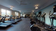 This fitness center offers a comprehensive and thoughtfully organized workout environment spread across a single floor. The space is divided into distinct functional zones, each catering to a different aspect of physical training. The layout is generously proportioned, providing ample room between equipment for comfortable use.

The cardio zone features a row of modern treadmills equipped with individual digital displays, alongside a lineup of elliptical trainers positioned along a light teal accent wall. Ceiling fans and pendant lighting fixtures are distributed throughout, ensuring consistent air circulation and illumination across the entire floor.

A dedicated strength training area includes a multi-station cable machine, a squat/power rack with weight plates, and adjustable benches, supported by wall-mounted mirrors that enhance the sense of space. Flat-screen televisions are mounted along the walls for entertainment during workouts.

Additional equipment includes stationary bikes, rowing machines, and a functional training rack stocked with medicine balls and stability balls in varying sizes, ideal for dynamic and flexibility-focused workouts.

Natural light enters through several windows positioned along the perimeter walls, offering views of the surrounding exterior. The flooring throughout is durable rubber matting, suitable for high-traffic fitness use. The overall ambiance is clean, modern, and well-maintained.