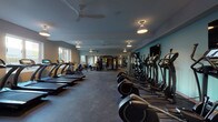 This fitness center offers a comprehensive and thoughtfully organized workout environment spread across a single floor. The space is divided into distinct functional zones, each catering to a different aspect of physical training. The layout is generously proportioned, providing ample room between equipment for comfortable use.

The cardio zone features a row of modern treadmills equipped with individual digital displays, alongside a lineup of elliptical trainers positioned along a light teal accent wall. Ceiling fans and pendant lighting fixtures are distributed throughout, ensuring consistent air circulation and illumination across the entire floor.

A dedicated strength training area includes a multi-station cable machine, a squat/power rack with weight plates, and adjustable benches, supported by wall-mounted mirrors that enhance the sense of space. Flat-screen televisions are mounted along the walls for entertainment during workouts.

Additional equipment includes stationary bikes, rowing machines, and a functional training rack stocked with medicine balls and stability balls in varying sizes, ideal for dynamic and flexibility-focused workouts.

Natural light enters through several windows positioned along the perimeter walls, offering views of the surrounding exterior. The flooring throughout is durable rubber matting, suitable for high-traffic fitness use. The overall ambiance is clean, modern, and well-maintained.