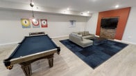 This single-floor recreation space is thoughtfully designed to balance active entertainment with comfortable lounging. The room features light hardwood-style flooring throughout, which provides a clean, contemporary foundation. A full-size pool table with a rich wood frame and deep blue felt dominates one side of the room, positioned beneath a pendant light that adds a warm, focused glow. Vintage-style number signs on the wall add a playful, casual character to the space.

On the opposite side, a modern gray sectional sofa is arranged atop a large navy area rug, creating a defined lounge zone ideal for gathering. A sleek coffee table anchors the seating area. An accent wall painted in a bold burnt orange introduces a striking pop of color, complemented by a stacked stone feature wall that adds texture and warmth near what appears to be a media area. Recessed lighting throughout ensures the space is well-lit and functional. The overall ambiance is energetic yet inviting, making it well-suited for hosting guests or casual everyday use.