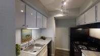 This single-floor residence offers a thoughtfully designed layout that maximizes functionality and comfort. The kitchen is a standout feature, showcasing white upper cabinetry that provides ample storage space, complemented by speckled granite-look countertops that add a clean, modern aesthetic. A stainless steel double-basin sink sits beneath the cabinetry, and the space is equipped with black appliances, including a full-size refrigerator and a range, delivering a classic contrast against the lighter tones of the room. Track lighting mounted on the ceiling ensures the kitchen is well-lit, while a ceiling vent contributes to efficient climate control throughout the space. The flooring features wood-look planks that run throughout, adding warmth and continuity to the interior. The walls are finished in a soft, neutral gray tone with a lightly textured surface, creating a calm and cohesive ambiance. A doorway off the kitchen leads seamlessly to additional areas of the residence, reinforcing the practical and connected flow of the floor plan. Overall, the space presents a comfortable and efficiently organized living environment with tasteful, functional design choices.