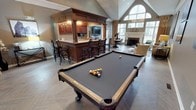 This single-floor clubhouse is designed to serve as a premier social and recreational hub. The open-concept layout accommodates multiple activity zones that flow seamlessly into one another, creating an inviting and versatile environment. A full-length billiards table anchors the foreground, set atop herringbone-patterned hardwood flooring that adds a refined, classic touch throughout the space.

A substantial dark wood bar with overhead cabinetry, built-in lighting, and bar stool seating serves as the room's central architectural feature. The bar includes a mounted flat-screen television, making it well-suited for gatherings and events. Adjacent to the bar, a dining area with table and chair seating provides a comfortable spot for more formal socializing.

Toward the rear of the space, a cozy lounge area surrounds a traditional fireplace framed by elegant millwork, offering a warm retreat. Tall vaulted ceilings and a large arched window flood the space with natural light, enhancing the open, airy ambiance. The combination of rich wood finishes, neutral tones, and thoughtful furnishings gives the clubhouse a warm yet polished character throughout.