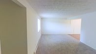 This two-floor space presents a straightforward and functional layout designed for comfortable everyday living. The interior is finished with a neutral color palette throughout, featuring crisp white walls that lend a bright, open feel to each room. The main living area is generously sized, with plush wall-to-wall carpeting underfoot that adds warmth and comfort to the space. A textured ceiling adds subtle visual depth overhead, while a baseboard heating system runs along the perimeter, ensuring efficient climate control throughout the colder months. Natural light filters in through a window, brightening the room and contributing to the overall airy ambiance. The transition between carpeted and hard-surface flooring is visible toward the adjoining area, hinting at a practical delineation between living and functional zones. The open layout offers flexibility for a variety of furniture arrangements and uses. Spanning two floors, the space provides ample room across multiple levels, allowing for a clear separation between living and private areas. The clean, uncluttered interior makes it easy to envision and customize the space to suit a range of needs and personal styles.