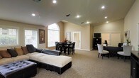 This single-floor space showcases an open-plan layout that seamlessly integrates multiple functional zones within one expansive room. The vaulted ceiling adds a sense of grandeur, drawing the eye upward while recessed lighting ensures consistent, well-distributed illumination throughout. Warm beige walls complement the neutral-toned carpet underfoot, creating a cohesive and inviting atmosphere.

The lounge area, positioned in the foreground, features a substantial dark-framed sectional sofa paired with a large ottoman, offering ample seating for relaxation or social gatherings. Toward the middle of the room, a dining area accommodates a dark wood table surrounded by matching chairs, positioned near a large arched window that floods the space with natural light. Decorative framed artwork adorns the walls nearby, adding visual interest.

Toward the rear, a dedicated workspace nook houses a computer workstation accompanied by a white accent chair, alongside a dark media console with a mounted flat-screen television. A secondary window in this area further enhances the natural brightness of the room. Overall, the space balances comfort, functionality, and an open, airy character.