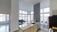 This single-floor space showcases a contemporary open-concept design that seamlessly integrates the kitchen, dining, and living areas into one cohesive environment. The kitchen serves as a central hub, anchored by a large island with a polished granite countertop that provides ample workspace and casual seating. Stainless steel appliances, including a dishwasher, complement the clean-lined cabinetry finished in a neutral tone, contributing to the space's modern aesthetic.

A striking floor-to-ceiling gray accent wall adds architectural depth and contrast against the predominantly white walls and ceiling. The living area benefits from dramatic ceiling height, enhancing the sense of openness and grandeur. A generous arrangement of large windows wraps around the space, flooding the interior with natural light and offering views of the surrounding area.

The flooring throughout reflects the light beautifully, reinforcing the bright, airy atmosphere. A dining area with contemporary transparent seating sits adjacent to the kitchen, maintaining visual flow. Overall, the space conveys a clean, minimalist character defined by thoughtful design and an abundance of light.