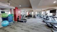 This fitness center offers a thoughtfully designed workout environment spread across a single floor. The space is anchored by bold red accent walls that contrast with neutral tones throughout, creating an energizing atmosphere suited for exercise. The interior features patterned carpet flooring and a coffered ceiling with recessed lighting, contributing to a clean and polished aesthetic.

The strength training area is well-stocked, featuring a cable machine, a full rack of dumbbells arranged on tiered shelving, a weight bench, and various medicine balls and free weights stored along the walls. This section provides ample room to move freely between equipment. The cardio zone, visible toward the rear of the space, is lined with treadmills and elliptical machines, all neatly arranged for easy access.

Wall-mounted mirrors run along one side of the fitness center, visually expanding the space and allowing users to monitor their form during workouts. Soft wall sconces provide supplemental lighting alongside the recessed ceiling fixtures, ensuring the entire area is well-lit. The overall layout is open and uncluttered, offering a functional and welcoming environment suitable for a wide range of fitness routines.