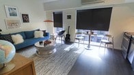 This single-floor space showcases a thoughtfully designed living area that balances modern aesthetics with personal character. Large floor-to-ceiling windows line the exterior wall, flooding the interior with natural light while offering views of the surrounding urban environment. Black roller blinds provide flexible light control and privacy when needed. An overhead wall-mounted air conditioning unit ensures year-round climate comfort.

The living area is anchored by a vibrant blue patterned sofa adorned with decorative throw pillows, paired with a distinctive hammered silver circular coffee table that serves as a striking focal point. A textured grey area rug defines the seating zone atop sleek dark flooring. A modern orange arc floor lamp adds a warm, artistic accent to the corner.

Near the windows, a casual high-top bistro table with bar-height chairs creates a secondary seating or dining nook, bathed in natural light. An acoustic guitar leaning against the wall introduces a creative, lived-in charm. A small framed artwork on the white wall adds a subtle decorative touch. Overall, the space presents a clean, contemporary urban character with eclectic personality.