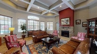 This inviting single-floor space makes a strong first impression with its dramatic coffered ceilings, which lend an air of architectural refinement and grandeur to the main gathering area. The soaring ceiling height draws the eye upward while recessed lighting and an abundance of large, arched windows flood the room with natural light and offer views of the surrounding exterior.

The focal point of the main lounge area is a stately stone fireplace set against a bold, deep-red accent wall, creating a warm and visually striking contrast to the neutral tones throughout the rest of the space. Comfortable leather sofas and ornately upholstered armchairs are arranged around a dark wood coffee table, all anchored by a richly patterned area rug over tile flooring. A wooden bookcase and framed artwork contribute to the space's refined, classic character.

An elegant arched passageway leads into a fully equipped kitchen, where warm wood cabinetry and stone-toned granite countertops define the workspace. A generously sized wraparound breakfast bar with cane-back bar seating provides a practical and social gathering point. Stainless steel appliances and ample cabinet storage complete the functional kitchen layout. Throughout the space, consistent tile flooring, coordinated warm color tones, and thoughtful furnishings create a cohesive, welcoming atmosphere suited for community gathering and entertainment.