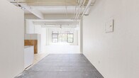 Located in the heart of Downtown Los Angeles on S Spring Street, this one-bedroom, one-bathroom live/work loft offers an open, versatile layout with a distinctly urban character. The space is defined by its exposed ceiling with visible ductwork and piping — a hallmark of the loft-style aesthetic — paired with crisp white walls throughout that create a bright, clean canvas. Large black-framed windows line the main living area, flooding the interior with generous natural light and offering views of surrounding city greenery and architecture. The floors feature a mix of painted white wood planks in the bedroom and living areas, contrasted by dark tile in portions of the main space, adding visual texture. A built-in shelving unit with lower cabinetry provides practical storage in the living area. The spacious bedroom continues the airy, light-filled theme with beamed ceilings, white plank flooring, open closet shelving, and a window overlooking the outdoors. The kitchen and dining area offer a functional layout with wood-accented cabinetry. The bathroom includes a combined tub and shower with glass enclosure, a wall-mounted sink, and dark flooring. This loft's industrial-meets-minimalist character suits both residential and creative studio use.