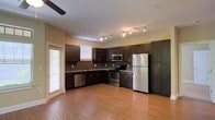 This single-floor space is thoughtfully designed to blend functionality with modern style. The kitchen serves as a central highlight, featuring rich, dark espresso cabinetry that extends floor to ceiling, providing generous storage throughout. A full suite of stainless steel appliances — including a refrigerator, electric range, built-in microwave, and dishwasher — offers a sleek, contemporary finish. Track lighting runs along the ceiling, casting bright, even illumination across the workspace, while a ceiling fan adds comfort to the open area. Warm-toned hardwood flooring flows seamlessly through the space, creating a cohesive and inviting atmosphere. Natural light filters in through multiple windows fitted with horizontal blinds, enhancing the airy feel of the interior. The kitchen layout is designed in an L-shape, maximizing counter space and workflow efficiency. The neutral wall tones complement the dark cabinetry and light flooring, resulting in a balanced and visually appealing contrast. Overall, the space presents a clean, modern aesthetic with practical features well-suited for everyday living.