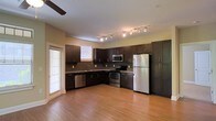 This single-floor space is thoughtfully designed to blend functionality with modern style. The kitchen serves as a central highlight, featuring rich, dark espresso cabinetry that extends floor to ceiling, providing generous storage throughout. A full suite of stainless steel appliances — including a refrigerator, electric range, built-in microwave, and dishwasher — offers a sleek, contemporary finish. Track lighting runs along the ceiling, casting bright, even illumination across the workspace, while a ceiling fan adds comfort to the open area. Warm-toned hardwood flooring flows seamlessly through the space, creating a cohesive and inviting atmosphere. Natural light filters in through multiple windows fitted with horizontal blinds, enhancing the airy feel of the interior. The kitchen layout is designed in an L-shape, maximizing counter space and workflow efficiency. The neutral wall tones complement the dark cabinetry and light flooring, resulting in a balanced and visually appealing contrast. Overall, the space presents a clean, modern aesthetic with practical features well-suited for everyday living.