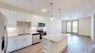 This well-appointed single-floor unit showcases a clean, contemporary design with an open-concept layout that maximizes both functionality and visual appeal. The kitchen serves as the centerpiece of the space, featuring crisp white cabinetry, a subway tile backsplash, and a full suite of stainless steel appliances including a range, microwave, dishwasher, and refrigerator. A central island with a speckled granite countertop provides additional prep space and open shelving below, while two stylish pendant lights hang overhead, adding a refined touch. Light hardwood-style flooring extends seamlessly throughout the unit, creating a warm and cohesive feel. The living area flows naturally from the kitchen, benefiting from large windows and glass double doors that invite abundant natural light and provide views of the surrounding exterior. A ceiling fan adds both comfort and a finishing detail to the space. The bedroom is generously proportioned, featuring the same hardwood-style flooring, a ceiling fan, and a sizable window that frames an exterior view. The bathroom is thoughtfully designed with white cabinetry, granite countertops, a framed mirror, and a full tub-shower combination, all finished in a clean, neutral palette. Overall, the unit presents a bright, modern aesthetic with quality finishes throughout.