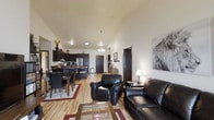 This single-floor residence offers an open-concept layout that brings together the living, dining, and kitchen areas in one cohesive and functional space. The living area is anchored by rich dark leather seating, including a sofa and armchair, arranged around a glass-top coffee table, creating an inviting space for relaxation. A large decorative wall piece adds a bold artistic touch to the room.

The dining area flows naturally from the living space, featuring a dark wood dining table with matching chairs, bordered by a patterned area rug. The kitchen is outfitted with dark cabinetry, stainless steel appliances including a refrigerator, and ample counter space, making it both stylish and practical.

Throughout the main living areas, warm-toned hardwood-style flooring ties the spaces together and contributes to the overall sense of openness. Vaulted ceilings add a sense of height and airiness to the layout, while a combination of overhead lighting and a chandelier-style pendant light above the dining area provides warm, layered illumination. A central hallway connects the living areas to additional rooms, providing a clear and organized flow throughout the space.