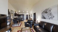 This single-floor residence offers an open-concept layout that brings together the living, dining, and kitchen areas in one cohesive and functional space. The living area is anchored by rich dark leather seating, including a sofa and armchair, arranged around a glass-top coffee table, creating an inviting space for relaxation. A large decorative wall piece adds a bold artistic touch to the room.

The dining area flows naturally from the living space, featuring a dark wood dining table with matching chairs, bordered by a patterned area rug. The kitchen is outfitted with dark cabinetry, stainless steel appliances including a refrigerator, and ample counter space, making it both stylish and practical.

Throughout the main living areas, warm-toned hardwood-style flooring ties the spaces together and contributes to the overall sense of openness. Vaulted ceilings add a sense of height and airiness to the layout, while a combination of overhead lighting and a chandelier-style pendant light above the dining area provides warm, layered illumination. A central hallway connects the living areas to additional rooms, providing a clear and organized flow throughout the space.