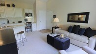 Located at 4130 Times Square Blvd in Dublin, Ohio, this single-floor residence offers a thoughtfully designed open-concept layout that maximizes both comfort and functionality. The living area is bright and inviting, anchored by plush carpeting, neutral wall tones, and soaring ceilings that contribute to an airy, spacious atmosphere. Large windows dressed with elegant draping panels flood the space with natural light and provide direct access to a private balcony, offering pleasant views of the surrounding greenery.

The living room is generously proportioned, accommodating a comfortable seating arrangement complemented by dark-toned accent furniture and ottomans that add a sophisticated contrast to the light palette. A dedicated built-in desk nook with cabinetry provides a practical workspace seamlessly integrated into the open floor plan. Adjacent to this, a breakfast bar with counter seating connects the living space to the kitchen area, ideal for casual dining or entertaining.

The bedroom is visible from the main living area and continues the cohesive design aesthetic, while a dark wood armoire provides stylish storage. The entryway features hardwood flooring, creating a clean transition into the carpeted living space. Overall, the residence presents a refined, comfortable environment with a balanced blend of practicality and style.