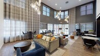 This single-floor amenity space is designed with a contemporary aesthetic that balances comfort and functionality. The centerpiece is a dramatic double-height lounge area featuring soaring ceilings, floor-to-ceiling patterned drapery, and an abundance of natural light streaming through large windows that offer views of the surrounding neighborhood. Clusters of globe pendant chandeliers cast a warm glow over an eclectic mix of tufted sofas, sectionals, accent chairs, and ottomans in neutral tones accented with pops of amber and navy.

A dedicated coworking zone is integrated seamlessly into the lounge, offering built-in desk stations with task seating and a printer, alongside a recessed nook furnished with booth-style seating and a world map art piece — ideal for focused work or small group meetings.

The kitchenette is fully outfitted with quartz countertops, wood-grain cabinetry, a commercial-grade beverage machine, microwave, refrigerator, and sink. A peninsula with gold-accented bar stools provides casual counter seating. A leasing or reception counter with additional quartz surfaces and organized display items completes the space.

Throughout, warm hardwood-style flooring unifies the zones, while recessed lighting and statement pendant fixtures enhance the modern, welcoming ambiance.
