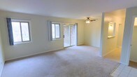 This single-floor space features a spacious, open living area finished with wall-to-wall carpeting in a neutral tone, creating a clean and comfortable foundation. The room is bathed in natural light through a combination of a standard window and a set of sliding glass doors, which offer a view of an adjacent brick exterior and provide convenient access to an outdoor area. The sliding doors serve as a primary architectural feature of the room, drawing the eye and contributing to an airy, open feel. A ceiling fan with a built-in light fixture is centrally mounted, ensuring year-round comfort and additional ambient lighting. The walls are painted in a soft, warm white, complementing the light carpeting and contributing to the overall brightness of the space. Structural columns are present and subtly define areas within the open layout without interrupting the sense of flow. White baseboards trim the perimeter of the room, adding a clean, finished look. The overall ambiance is calm and understated, offering a versatile, move-in-ready environment well-suited for a variety of furniture arrangements and personal styles.