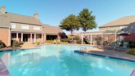 This expansive outdoor pool area serves as a central amenity within a well-maintained brick residential community. The centerpiece is a generously sized swimming pool with crystal-clear blue water, accessible via built-in handrail entries and framed by a broad surrounding deck ideal for lounging and socializing under open skies.

The pool deck transitions into a spacious sun-bathing area furnished with numerous cushioned chaise lounges, providing ample space for residents to relax and soak up natural light. A striking wooden pergola anchors one section of the outdoor area, offering shaded seating with stylish upholstered furniture — a comfortable retreat from the sun. Additional shaded dining options are available through umbrella-topped tables and chairs positioned throughout the space.

Mature trees border the perimeter, adding natural shade and a sense of privacy, while manicured shrubs and landscaped garden beds enhance the overall aesthetic. A decorative iron fence encloses the area, providing security without obstructing views of the surrounding multi-story brick buildings with balconies. A white lattice pergola near the pool adds an architectural accent that complements the community's traditional design. The entire space is open to the sky, bathed in abundant natural light, and designed to encourage an active, relaxed outdoor lifestyle.