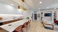 Located at 1630 Lewis Landing Ave in Wilmington, NC, this single-floor amenity space offers a refined blend of contemporary design and functional versatility. The open layout is anchored by a central lounge area featuring a curved white sectional sofa, cognac leather accent chairs, and a striking built-in wall unit with open wood shelving flanking a mounted television. Light hardwood flooring runs throughout, complemented by modern sputnik-style chandeliers that add an architectural touch overhead.

The kitchenette is a standout feature, showcasing a generous marble-topped island with seating for several, rich walnut cabinetry, a backlit marble tile backsplash, and a matte black sink fixture. A dramatic brass pendant light anchors the space with warm, sophisticated detail.

Multiple private meeting or lounge rooms provide quieter settings, each with distinct wallcoverings, plush seating, and large windows that draw in natural light and views of the surrounding greenery. A cozy window nook with built-in seating offers an inviting retreat. Dining areas with designer pendant lighting and round tables provide flexible gathering spots. The space also includes ADA-accessible restrooms with terrazzo tile flooring, ensuring inclusivity throughout.