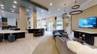 This sophisticated single-floor communal space is designed with an open-concept layout that seamlessly blends comfort and modern elegance. The lounge area is anchored by a pair of striking wrought-iron chandeliers adorned with exposed bulbs, casting warm ambient light throughout the room. Plush, tufted gray sofas and ottomans are arranged to encourage relaxed conversation, accented by cozy throws and sleek side tables that add a touch of warmth to the refined aesthetic.

A curved transition between hardwood-style flooring and soft carpeting subtly delineates the lounge from the surrounding areas, adding an architectural detail that enhances the flow of the space. Tall, square columns punctuate the room, contributing to its sense of structure while maintaining an airy, unobstructed feel.

Toward the rear, a well-appointed bar area with dark cabinetry and high counter seating provides a dedicated social hub, ideal for gatherings. Large, floor-to-ceiling windows and glass double doors flood the interior with natural light and offer views of the outdoor surroundings. Recessed ceiling lighting complements the chandeliers, ensuring the space is well-lit at all hours. Decorative wall clocks and carefully chosen greenery complete the polished, inviting atmosphere of this communal retreat.