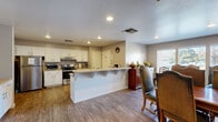 Located in Antioch, California, this single-story community space is thoughtfully designed to accommodate both everyday living and social gatherings. The layout flows seamlessly between functional and recreational areas, all unified by warm wood-look flooring throughout.

The kitchen is well-appointed with white shaker-style cabinetry, stainless steel appliances including a refrigerator, range, and microwave, and a central peninsula with bar seating. Generous counter space and ample storage make it well-suited for communal meal preparation. An emergency exit door adds a practical safety feature to this area.

Adjacent to the kitchen, the formal dining room offers an inviting atmosphere with a large wooden dining table and high-backed upholstered chairs that seat a sizable group. Large windows fitted with horizontal blinds line the walls, allowing abundant natural light to fill the room while providing views of the surrounding outdoor environment.

A graceful arched passageway leads into a spacious recreation room, which serves as a central gathering area. This room features a full-size pool table as a focal point, recessed and track lighting overhead, and an open layout ideal for leisure and socializing. A built-in bar counter and bookcase add character and functionality to the space. The textured ceilings and neutral wall tones carry throughout, lending the entire space a cohesive and welcoming ambiance.