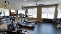 This fitness room offers a practical and comfortable environment for cardio-focused workouts. The space is outfitted with multiple Nautilus treadmills and an elliptical trainer, providing ample options for cardiovascular exercise. Rubber flooring covers the entire room, offering durability and support underfoot. Natural light flows generously through several large windows dressed with white horizontal blinds, creating a bright and energizing atmosphere. The warm, olive-toned walls contribute to a grounded, focused ambiance throughout the space. A ceiling fan positioned centrally overhead aids in air circulation, enhancing comfort during workouts. A wall-mounted flat-screen television is positioned at an ideal viewing angle, allowing users to stay entertained while exercising. Recessed lighting supplements the natural light, ensuring the room remains well-lit regardless of the time of day. The layout is thoughtfully arranged, with equipment positioned to maximize the available floor space while maintaining clear pathways. Overall, the fitness room is a functional and inviting space designed to support an active lifestyle.