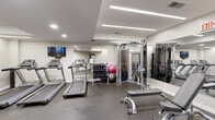 Located at 9 Municipal Dr in Fishers, Indiana, this fitness center offers a thoughtfully designed, single-floor exercise space packed with a comprehensive range of workout equipment. The gym features a modern aesthetic with crisp white walls, speckled rubber flooring for safety and durability, and a combination of recessed and linear LED ceiling lighting that creates a bright, energizing atmosphere.

The cardio section is well-appointed with multiple treadmills, elliptical machines, and a stationary cycling bike, catering to a variety of aerobic workout preferences. A wall-mounted flat-screen television accompanies the cardio area, providing entertainment during workouts. The strength training zone includes a multi-station cable machine, a dedicated weight bench, a full dumbbell rack with an array of free weights, and stability balls, offering versatile options for resistance training.

Full-length mirrors line portions of the walls, enhancing the sense of space while allowing users to monitor their form. The facility also includes practical amenities such as a water dispenser station and a sanitizing station near the entrance, ensuring a clean and comfortable environment. The space is accessed through a keyed entry door and also features an emergency exit, reflecting careful attention to safety and convenience throughout the design.