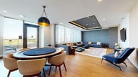 Located at 300 McRae Avenue in Ottawa's vibrant west end, this thoughtfully designed amenity floor spans a generous single level and comprises six distinct areas crafted for comfort and socializing. The interiors are defined by a sophisticated contemporary aesthetic, highlighted by striking coffered ceilings with integrated LED cove lighting, warm hardwood-style flooring, and carefully curated wallcoverings featuring bold geometric patterns in muted blue and gold tones. The main lounge area is a standout, offering expansive seating arrangements with plush sofas, layered area rugs, and floor-to-ceiling windows dressed in sheer drapery that flood the space with natural light. A dedicated games and dining zone features a round table with upholstered chairs beneath a dramatic black-and-gold pendant light. A quieter retreat room provides a more intimate setting with a sofa, patterned accent wall, and slatted wood paneling. The well-appointed corridor connecting the spaces is adorned with curated artwork and warm sconce lighting. Outside, a spacious tiled terrace wraps around the building, enclosed by glass-panel railings, and delivers unobstructed panoramic views of the Ottawa skyline and surrounding greenery — ideal for outdoor gatherings in any season.