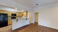 This single-story space is designed with an open-concept layout that seamlessly connects the kitchen and living area, creating a functional and welcoming environment. The kitchen is outfitted with light maple cabinetry offering generous storage, a neutral travertine tile backsplash, and granite countertops that extend to form a peninsula-style breakfast bar with corbel supports — a practical spot for casual dining or additional prep space. Black appliances, including a gas range and built-in microwave, provide a clean contrast against the lighter tones of the cabinetry and counters. Recessed lighting illuminates the kitchen efficiently. Crown molding runs along the ceiling throughout, adding a refined architectural detail that elevates the overall aesthetic. Rich, warm-toned hardwood flooring spans the open living area, contributing to a cohesive and inviting atmosphere. The white walls keep the space feeling bright and neutral. A doorway visible from the main area leads to additional rooms, suggesting a practical and well-connected floor plan. Overall, the space balances everyday functionality with clean, understated design details.